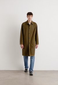 Brown waterproof coat with a collar, button closure, and two front pockets. Paired with light blue jeans and black shoes.