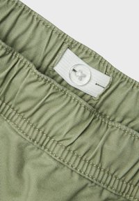 Ceinture élastique en tissu vert clair, avec une étiquette blanche et un bouton en plastique transparent. Détails subtils de texture et de couture.