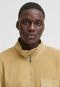 Fleece jacket in light yellow with a high collar, zip closure, and single chest pocket. Soft texture and minimalistic design.