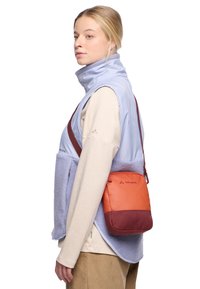 Gilet senza maniche blu chiaro e viola con texture in pile, abbinato a una maglietta a maniche lunghe color crema e a una borsa a tracolla arancione e borgogna.