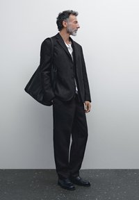 Black, tailored suit with a single-button jacket and straight-leg trousers. Crisp white shirt underneath. Black leather bag and shoes.