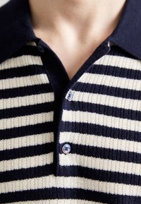 Detailní záběr na osobu, která má na sobě pletenou polo košili s námořnickou modrou a krémovou pruhovanou vzorováním, se dvěma modrými knoflíky zapnutými u límečku.