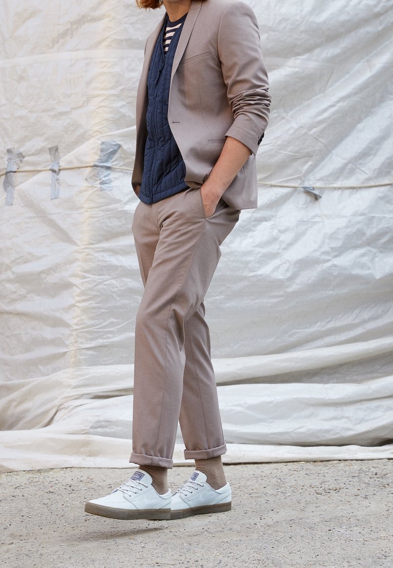 Personne portant un pantalon et une veste de costume beige avec un gilet matelassé bleu, une chemise rayée, des chaussettes beiges et des baskets blanches, debout devant un fond blanc.