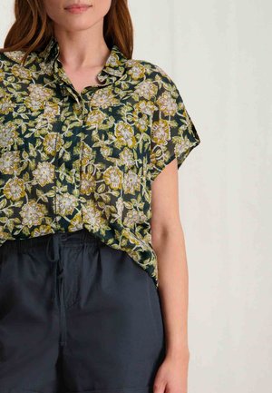 Korte mouwen blouse met bloemenpatroon in donkergroen, met witte en gele bloemen; lichtgewicht stof met een relaxte pasvorm. Zwarte shorts zichtbaar eronder.