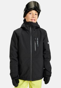 Młoda osoba ubrana w czarną zimową kurtkę, czarne rękawiczki, czapkę beanie w kolorze czarnym z goglami narciarskimi oraz jasne żółte spodnie narciarskie na jednolitym tle.