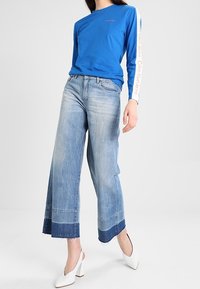 Blå långärmad skjorta med vit logotyp på ärmarna, kombinerad med vida jeans i blekt denim med fransade fållar och kontrasterande band.