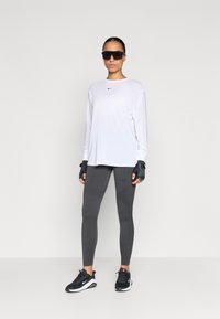 Biele tričko s dlhým rukávom značky Nike, šedé legíny s rebrovanou textúrou, čierne rukavice a čierne tenisky Nike. Veľké čierne slnečné okuliare.