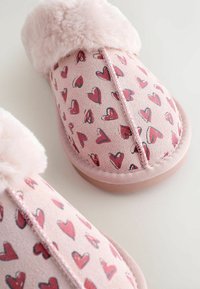 Lichtroze pantoffels met een zachte voering van bont. Het bovenwerk heeft een ontwerp met verschillende rode harten op een suède-achtige stof. Platte rubbersoal.