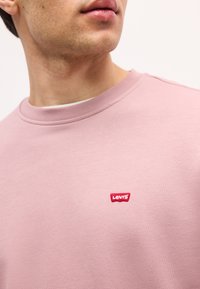 Νεαρός άνδρας που φοράει ένα ανοιχτό ροζ φούτερ με στρογγυλή λαιμόκοψη και ένα μικρό κόκκινο λογότυπο Levi's κεντημένο στο στήθος.