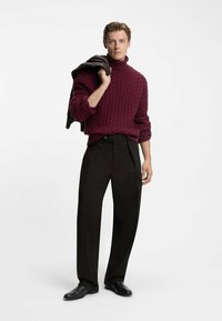 Maglione a collo alto lavorato a maglia di colore bordeaux con un motivo texture, abbinato a pantaloni neri a gamba larga e scarpe nere, giacca appoggiata sulla spalla.