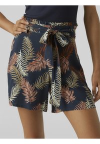 Žena v námořnických kraťasech s béžovým a rezavým listovým vzorem a uvázaným tkaninovým opaskem, který má na sobě černý top.