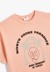 Kortærmet, ribbet t-shirt i blød lyserød med sort og teal grafik: "NORTH SHORE PARADISE" med palmer og et kraniumdesign.