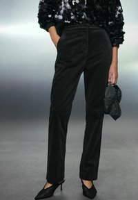 Schwarze Corduroyhose mit geraden Beinen und seitlichen Taschen. Kombiniert mit einem funkelnden schwarzen und silbernen Oberteil und spitzen schwarzen Absätzen.
