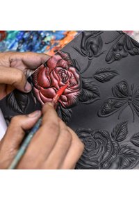 Rote Farbe wird von Hand auf eine strukturierte schwarze Oberfläche aufgetragen, die erhabene Rosen und Schmetterlinge zeigt, und dabei werden filigrane Details und Tiefe präsentiert.