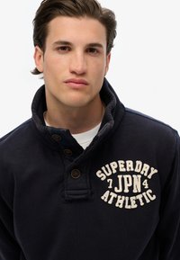 Granatowy sweter z bawełnianej mieszanki z wysokim kołnierzem, posiadający trzy brązowe guziki oraz białą haftowaną napis "SUPERDRY JPN 4 ATHLETIC."