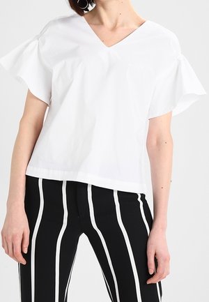 Personne portant un haut blanc à manches courtes en V avec des manches volantes et un pantalon noir à rayures blanches verticales.