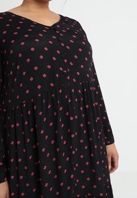 Robe noire à manches longues, décolleté en V et petit motif floral rose. Présente des détails de boutons et une jupe ample et froncée.