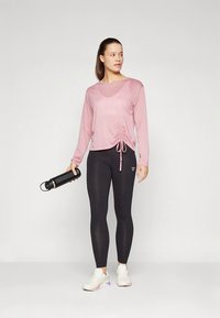 Pink langærmet top med sideløbning, stylet med sorte leggings og hvide træningssko. Modellen holder en sort vandflaske.