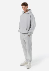 Sudadera con capucha gris claro, con un bolsillo frontal, pantalones de chándal a juego y zapatillas blancas; presenta un tejido cepillado y puños acanalados.