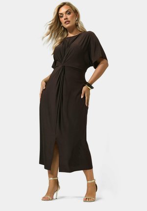 Femme aux longs cheveux blonds portant une robe midi noire avec un twist et une fente à l'avant, des talons hauts dorés, des boucles d'oreilles créoles et des bracelets, se tenant avec confiance.
