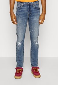 Blå denim jeans med en slitna design, med revor och färgfläckar. Matchas med röda skor med gula snören och tan-färgade sulor.