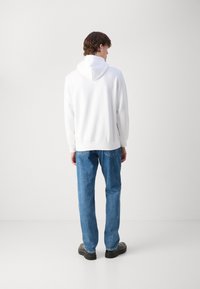 Weißer Kapuzenpullover mit Kängurutasche, verstellbarer Kapuze und gerippten Bündchen, kombiniert mit blauen Jeans und schwarzen Schuhen, von hinten gezeigt.