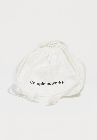 Borsa in cotone con cordoncino di chiusura, di colore bianco e finitura testurizzata. Presenta il testo nero "Completedworks" sulla parte anteriore. Forma compatta e rotonda.