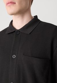 Črna polo majica z teksturo, ki ima ovratnik, dva črna gumba in žep na prsih. Tkanina ima rebrast vzorec.