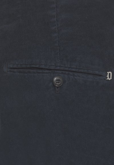 Primo piano di un tessuto in velluto blu scuro con una tasca orizzontale, bottone nero e piccolo emblema decorativo a forma di "D" argentato.