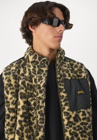 Umelá kožušinová vesta s leopardím vzorom v krémovej, hnedej a olivovej zelenej farbe, s čiernou nylonovou kapsou a zipsom. Čierne slnečné okuliare.