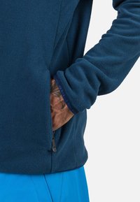 Giacca in fleece blu navy con tasca con zip e tessuto texturizzato. I polsini presentano un bordino blu navy a contrasto, mentre la giacca è indossata sopra pantaloni blu.