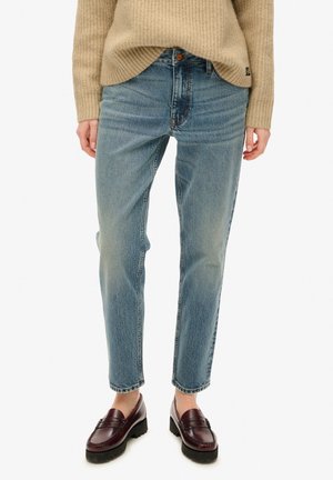 Persoon draagt lichtblauwe rechte spijkerbroek, beige gebreide trui en donkerbruine loafers met grove zolen, staand tegen een witte achtergrond.