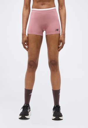 Isik, kellel on seljas roosad New Balance spordipüksid, tumedad sokid ja mustad jooksujalatsid, seisab valgel taustal.