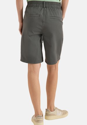Personne portant un short gris foncé longueur genou avec une taille élastique et des baskets beige, debout devant un fond uni.