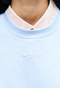 Κοντινό πλάνο ενός ατόμου που φοράει ανοιχτό ροζ πουκάμισο με γιακά κάτω από ένα ανοιχτό μπλε φούτερ Nike με λευκό κεντημένο λογότυπο Nike swoosh.