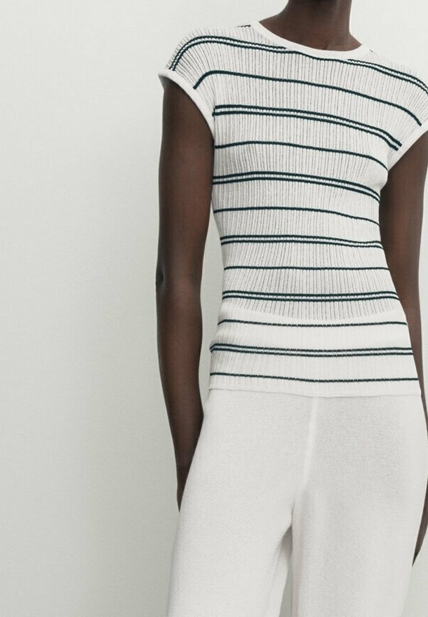 Personne portant un haut blanc sans manches côtelé avec des rayures horizontales noires et un pantalon blanc sur un fond clair uni.