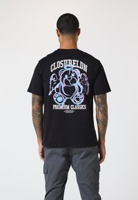 Czarna bawełniana koszulka z kolorową grafiką przedstawiającą niedźwiedzia z skrzydłami, z napisem "CLOSUBELDN PREMIUM CLASSICS" powyżej i poniżej wzoru.
