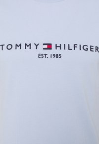 Logo Tommy Hilfiger avec un drapeau rouge, blanc et bleu marine entre les mots, et « EST. 1985 » imprimé en dessous sur un fond en tissu clair.