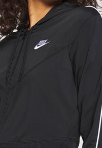 Zwarte rits-hoodie gemaakt van soepel materiaal, met een prominent paars Nike-logo, witte zijkanten en gestructureerde naden.