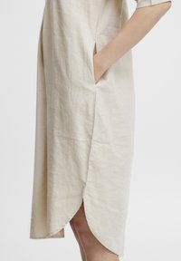 Robe en lin beige à manches courtes, avec poches latérales et un ourlet arrondi. Texture douce avec peu de plis. Design simple et droit.