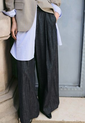 Persona con pantalones anchos de mezclilla oscura, camisa larga a rayas en capas, blazer beige y zapatos negros puntiagudos, de pie junto a superficies de piedra y metal.