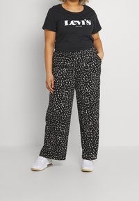 Pantalones negros con un patrón moteado blanco, diseño de pierna ancha, combinados con una camiseta negra de Levi's y zapatillas blancas. Look casual.
