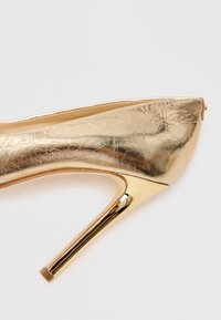 Gold metallic high-heeled shoe with a pointed toe and shiny stiletto heel on a light background.