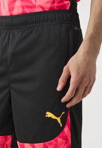 Pantaloni sportivi neri con logo Puma giallo, caratterizzati da una texture liscia e una vita elastica, abbinati a una maglietta a fantasia rosa.