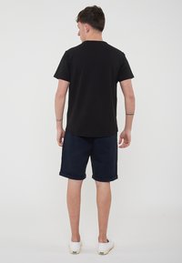 T-shirt nera a maniche corte con scollo rotondo, abbinata a pantaloni corti blu navy arrotolati. Modello con scarpe bianche slip-on.