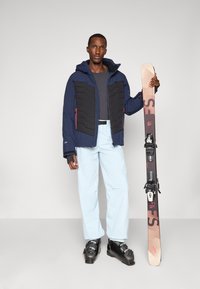 Marineblau und schwarze isolierte Jacke mit Kapuze, hellblaue Hose und schwarze Skischuhe. Hält einen braunen Ski mit schwarzen Bindungen, Text ist sichtbar.