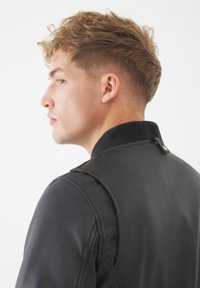 Chaqueta de cuero negra con un collar acanalado, que presenta una textura suave y un diseño elegante. Costuras visibles y un detalle de botón en el cuello.