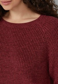 Burgunderfarbener Strickpullover mit gerippter Textur, weitem Rundhalsausschnitt und Raglanärmeln; der Stoff wirkt weich und warm.