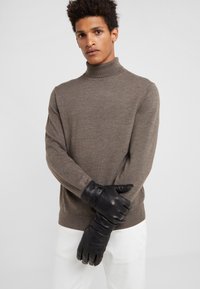 Brauner Rollkragenpullover aus weichem Material, kombiniert mit schwarzen Lederhandschuhen mit Riemen-Details, vor einem neutralen Hintergrund.