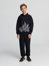 Felpa nera con cappuccio e motivo a fiamma bianca sull'orlo, abbinata a pantaloni della tuta neri e scarpe nere. Il tessuto sembra morbido e casual.